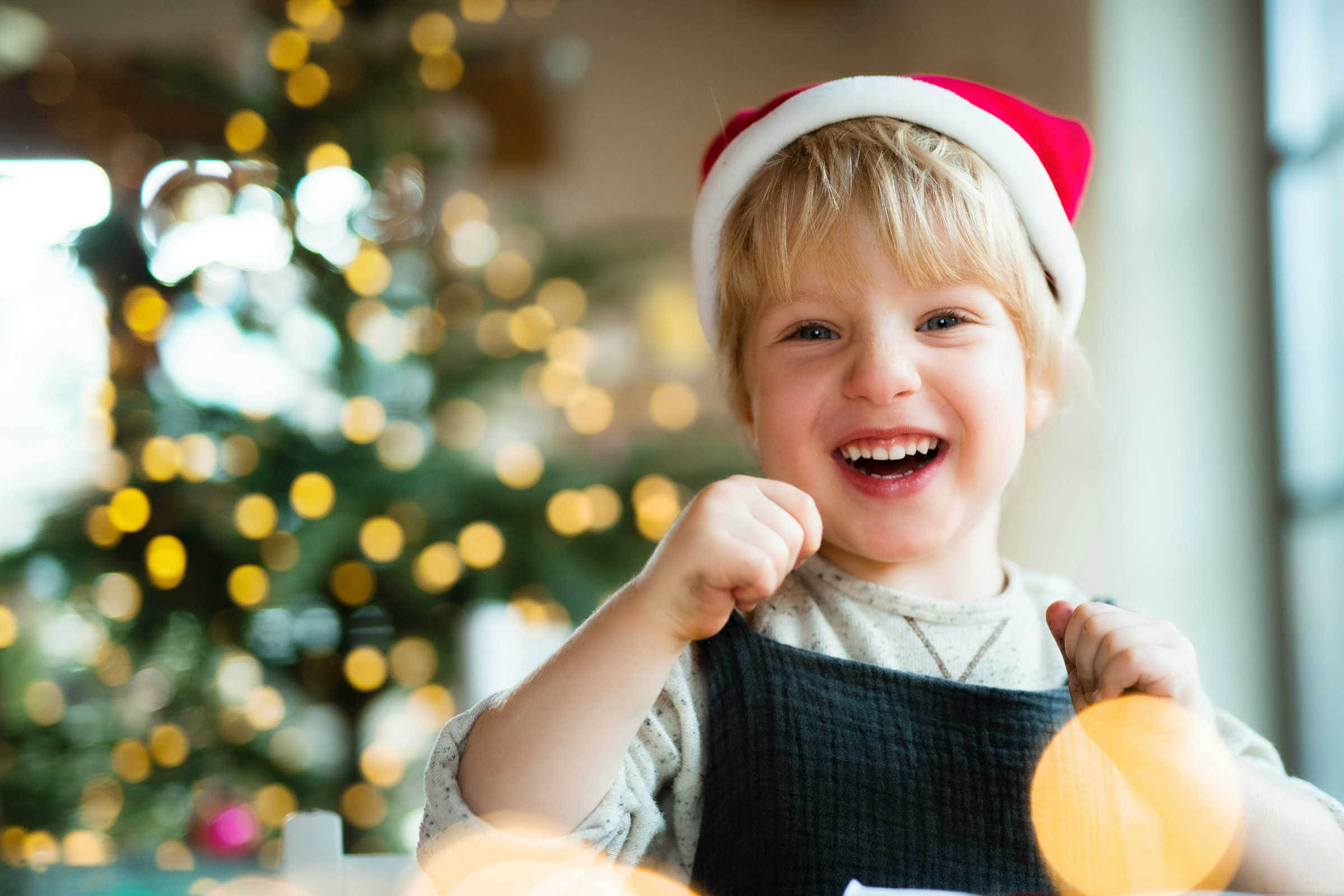 kleines Kind mit Weihnachtsmütze vor einem leuchtenden Weihnachtsbaum